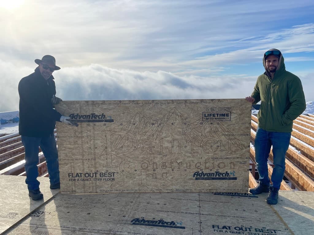 Two members of Rainier Construction on a jobsite above the clouds, holding sheathing marked with the company name
