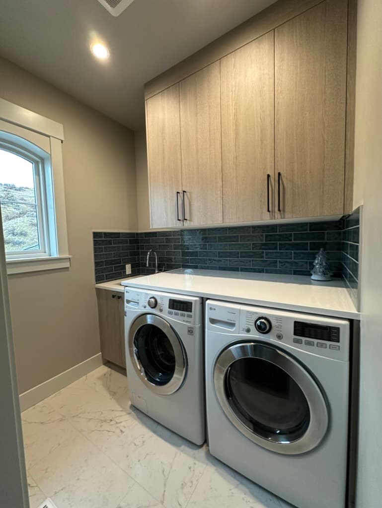Laundry room with light wood upper cabinets and a dark tile backsplash