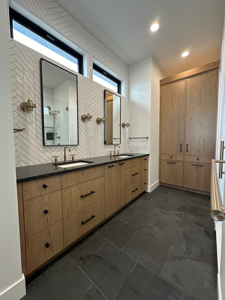 Primary bath with a light wood vanity, herringbone wall tile, and black counters